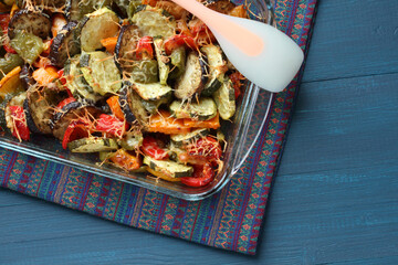 Vegetable casserole with zucchini, paprika, eggplant and cheese, baked in a glass form, which stands on a blue wooden table. View from above. Closeup