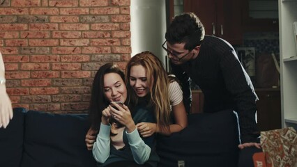 Girl sitting on couch looking at phone screen. Friends excited and chatting.Couple of friends come from behind, interested in activity and sharing news. Concept of internet, chatting, technology.