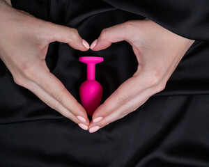 Woman making a heart shape with her palms around a pink butt plug on a black silk sheet. 