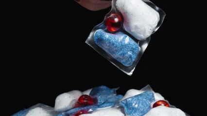 A woman's hand picks up one soluble packet with dishwasher powder. The background is black, and there are multiple capsules around.