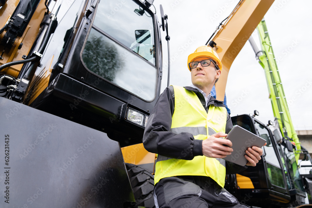 Poster engineer in a helmet with a digital tablet next to construction excavators