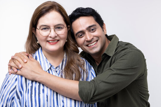 Portrait of smiling Indian mid adult man with hand on mature mother's shoulder standing together against white background
