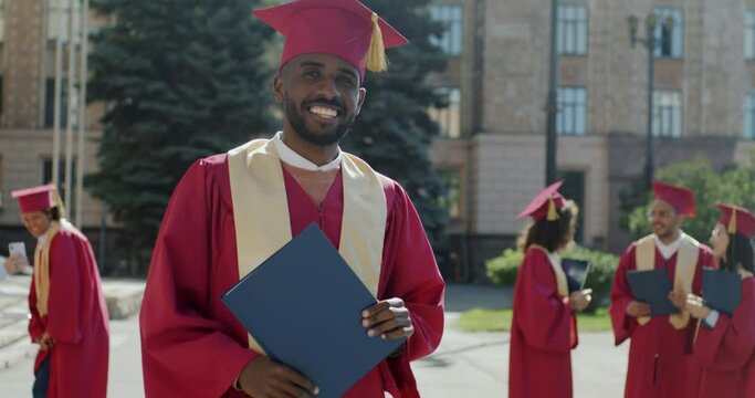 Slow motion portrait of successful African American man in graduation mantle and hat standing on campus holding diploma smiling looking at camera