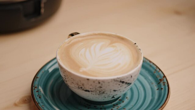 Female hand of the barista puts the finished beautiful cappuccino coffee in a cup on a saucer on the wooden table. Slow motion closeup shot with bokeh