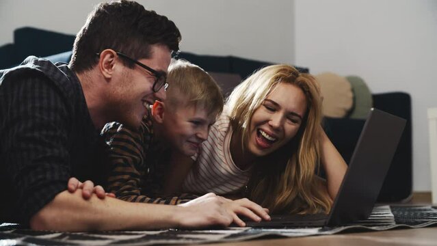 Smiling family looking at computer screen and enjoying watching funny social media video. Happy young parents and blond teenage son using laptop while lying on the floor. Concept of being at home.