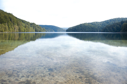 Krystalicznie czyste jezioro z gładką taflą pośród zalesionych górek w Plitvickim Parku Narodowym.