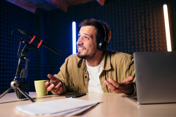 Young stylish man in mustard shirt with headphones gesturing at microphone and sharing story with...