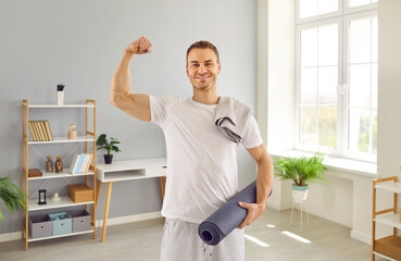 Portrait of happy fit male athlete after active fitness workout at home. Smiling handsome sporty man in white T shirt standing in living room, holding sports mat and showing strong biceps arm muscles