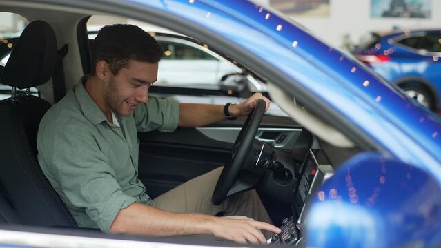 Smiling young man buyer sitting inside new car and checking all function of new model at showroom. Handsome male customer examining various auto during visit at dealership office, slow motion.
