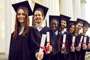 Happy confident graduate students standing in a row in black robes with diplomas in their hands...