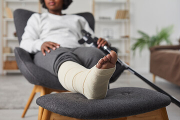 African american woman is sitting at home in armchair with broken bandaged leg lying on stool and...