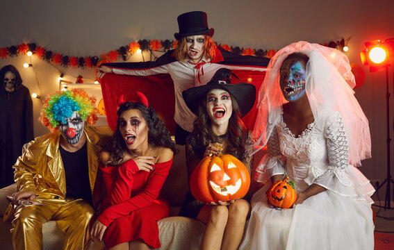 Portrait of group of adult creative friends in various spooky images at Halloween party. Multiracial friends sitting on sofa in room and having fun making eerily funny grimaces while looking at camera