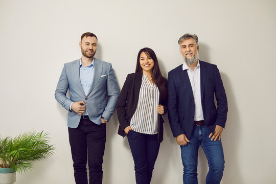 Team of young and mature business people at work. Group portrait of happy colleagues in office. Men and woman wearing smart casual clothes standing by white office wall, smiling, and looking at camera