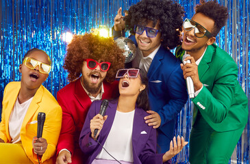 Happy multiracial friends wearing funny curly wigs and colourful disco suits singing karaoke in...