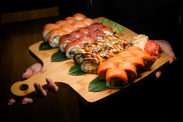 Woman's hand holds large wooden board of Philadelphia Sushi roll set with salmon, tuna, and eel