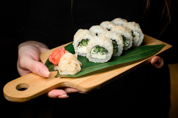 Woman's hand holds wooden board of California sushi rolls with Chuka alggae, cream cheese and sesame served decorated with green leaves and ginger