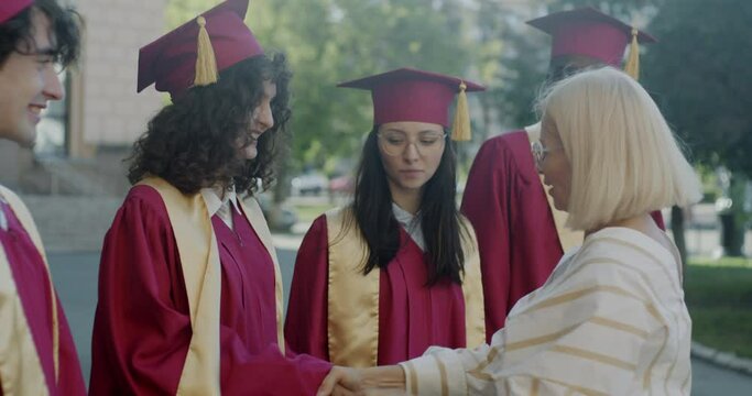 Happy dean congratulating multiethnic group of students shaking hands hugging young people in graduation gowns and caps. Education and successful youth concept.