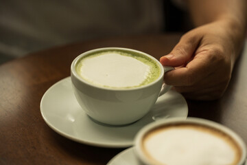 Closeup hand holding white cup of hot green tea latte and blurry hot coffee latte cup on wood background in restaurant.Best of menu in the coffee shop.Soft focus.