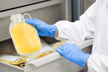 Doctor hand wearing blue gloves holding fresh frozen plasma bag in storage blood refrigerator at blood bank unit laboratory.Human plasma for covid-19 patients treatment