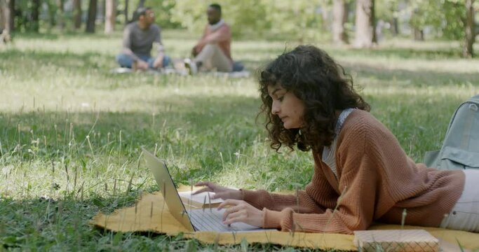 Young lady studying for exam in park working with laptop and reading textbook lying on blanket on lawn. Education and nature concept.