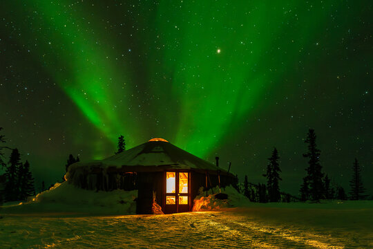 Nighttime image of the green northern lights or aurora borealis rising from behind a snow covered yurt with firelight coming from inside the yurt.