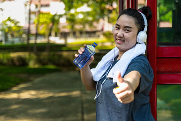 Fat woman with headphones drinking bottle water and making thumbs up after doing exercise in park, Workout running in nature