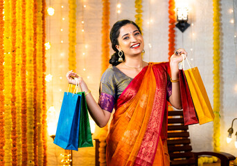 Happy young indian girl in saree with shopping bags in hand looking at camera at home - concept of...
