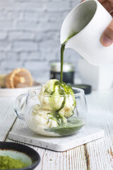 A glass of vanilla ice-cream while hand pouring matcha green tea on the top of it or it's called matcha affogato. With it's powder in a scene and bamboo chasen whisk in background 