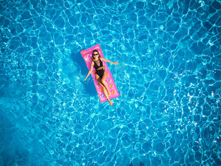 Fit pretty girl in bikini chilling on inflatable pink mattress in swimming pool. Slim hot woman in swimwear tanning. Female relaxing on float in blue water at luxury resort. Aerial, view from above.