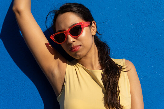 Happy young ethnic woman with sunglasses standing in light against blue wall