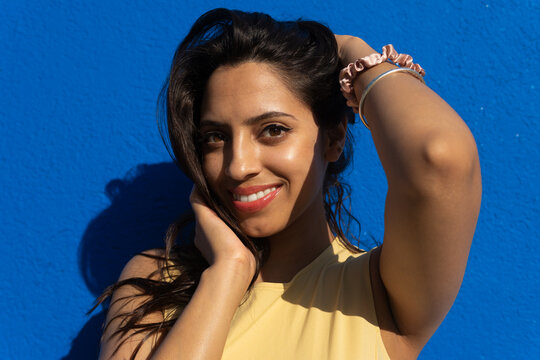 Smiling ethnic woman touching hair and standing against blue wall