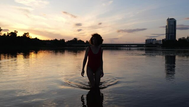 A woman in a red swimsuit comes out of the water in the evening at sunset. Woman relax beach slow motion