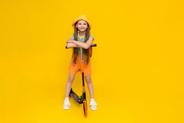 A full-length happy, cheerful young girl in a hat and shorts riding a two-wheeled scooter. Children's active city recreation. Bright yellow isolated background.