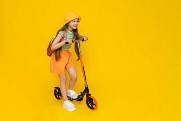 A teenage girl rides a scooter in shorts, a panama hat and with a backpack. Summer sports and active recreation for children. Yellow isolated background. © Юлия Дьякова