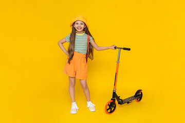 A cheerful young girl in a hat and shorts is going to ride a two-wheeled scooter. Children's active city recreation. Yellow isolated background.