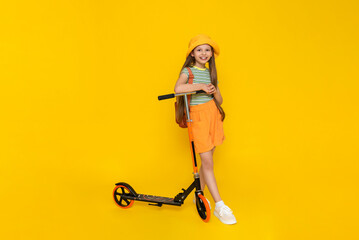 A full-length young girl in a hat and shorts is standing next to a scooter and enjoying a summer vacation. Active city recreation for children. Yellow isolated background.