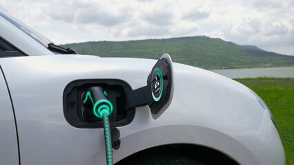 Electric car plug in with charging station, recharge battery by EV charger cable display smart digital battery status hologram with nature background. Futuristic green energy infrastructure.Peruse