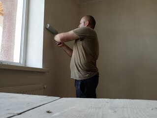 A middle-aged woman paints the walls in a room with the help of a roller. Repair in the room, stage of painting the walls.