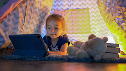 Happy cute little girl having fun playing in teepee tent. Child using digital tablet watching cartoons or playing computer games lying in kid tent at home. © Денис Максимчук