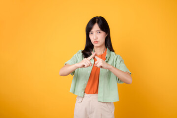 denial with a young Asian woman 30s, elegantly clad in orange shirt and green jumper. Her cross hand gesture, isolated on vibrant yellow background, represents concept of refusal and disagreement.
