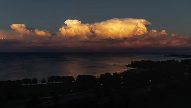 Sunset time lapse of airplanes emerging from white yellow and pink cumulonimbus clouds billowing up over Lake Michigan as swimmers take one last dip and a sailboat returns home as darkness sets in.