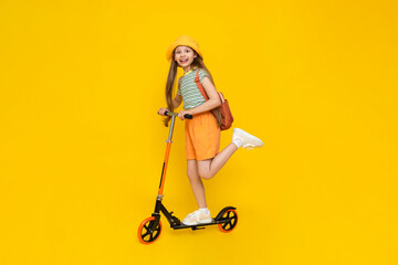 A child rides a scooter. A full-length side view of a beautiful cheerful dreamy young girl. Bright yellow isolated background.