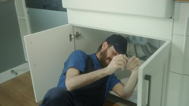 Young Hippie Plumber with Long Hair, Bearded, Hat-Wearing, Skillfully Repairs Sink Underneath