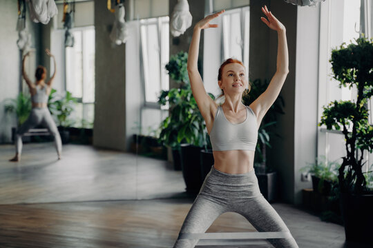 Slender redhead fitness instructor enjoys squats with pilates resistance band in gym, hands raised overhead. Workout joyfully amid mirrored walls.