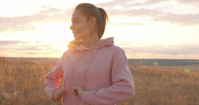 beautiful fit lady is wearing pink hoodie hood looking at side at sunset in contemplation, thinking, after jogging, sport training Dreamy sportswoman has rest, standing in field, stretching arms