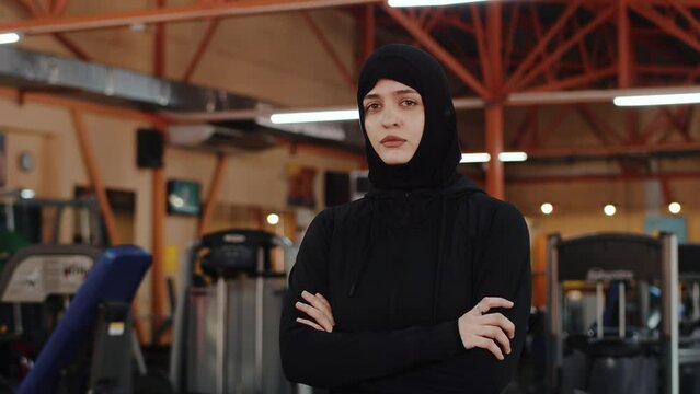 Young muslim female athlete in a hijab crossing her arms on her chest and looking at the camera with a confident face. Sportswoman in black sportswear before the training in an indoor gym