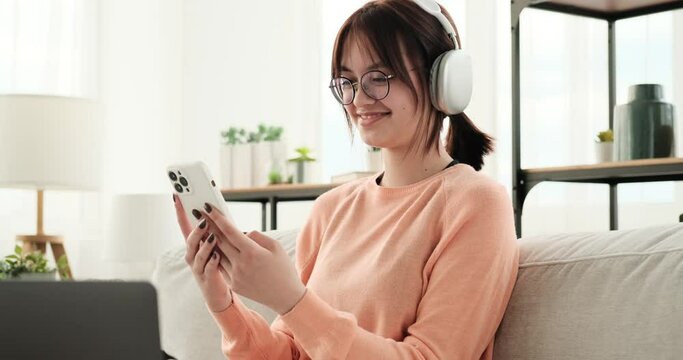 Seated comfortably on the couch, a smiling caucasian teenage girl enjoys using her phone while wearing headphones. With a contented expression, she engages in various activities on her phone.