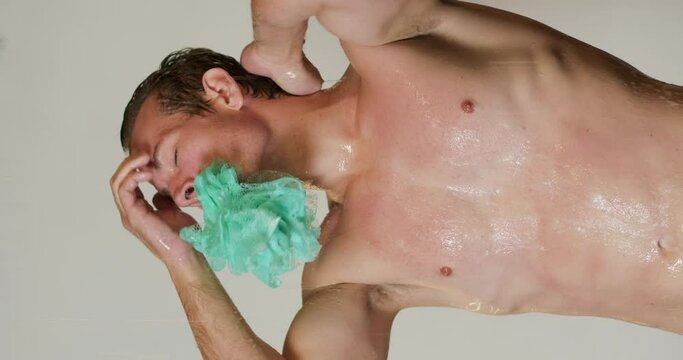 hot sexy young slim muscular man with sponge in mouth having morning water procedures standing under running water at home Slow motion attractive guy enjoying a hygienic shower, preparing for date