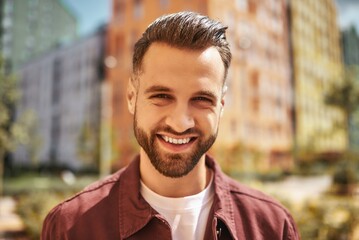 Hello Portrait of happy attractive man with stubble smiling and looking at camera while standing on the street