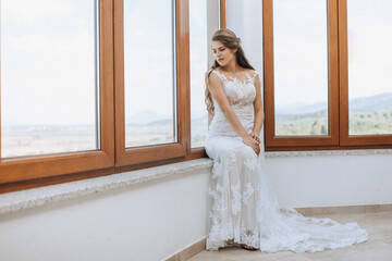 the bride in an evening dress sits on the windowsill against the background of large windows....